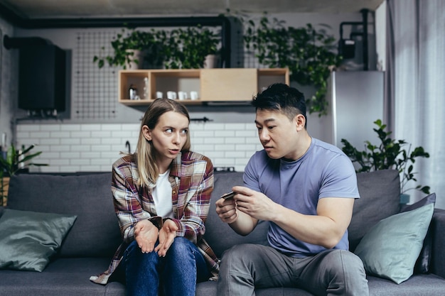 Asian man quarrels with his wife accuses him of squandering family money family quarrel couple sitting on sofa husband points to reduction of credit card account