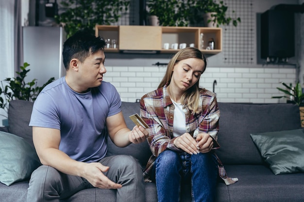 asian man quarrels with his wife accuses him of squandering family money family quarrel couple sitting on sofa husband points to reduction of credit card account
