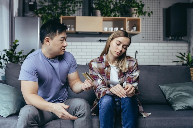 Photo asian man quarrels with his wife accuses him of squandering family money family quarrel couple sitting on sofa husband points to reduction of credit card account