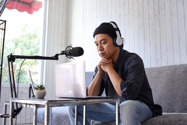 Asian man recording a podcast with headphones, microphone and script at home studio