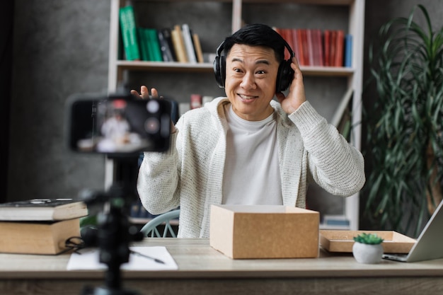 Asian man recording video on phone camera while unpacking box with new wireless headphones