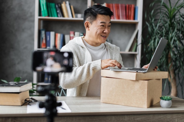 Asian man recording video on phone camera while unpacking box with new wireless laptop