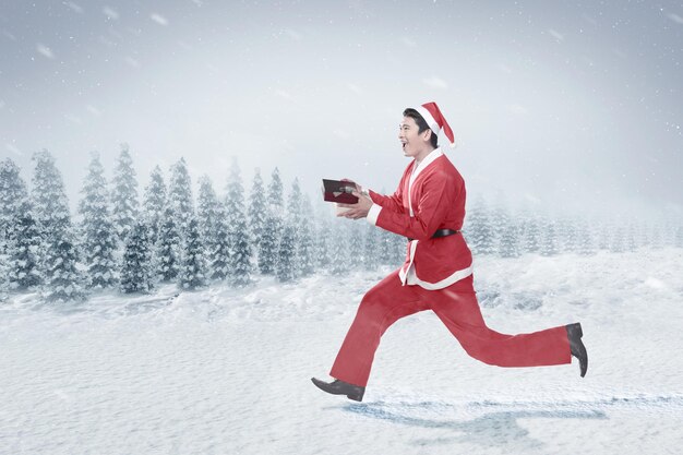 Asian man in Santa costume running while holding gift box on the snowfield with snowfall background. Merry Christmas