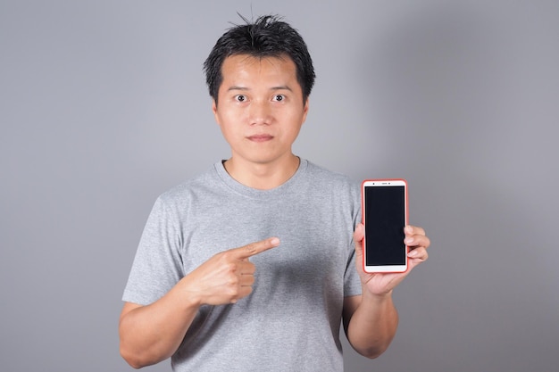 Uomo asiatico che mostra il telefono cellulare o lo smart phone dello schermo in bianco sullo schermo nero del fondo grigio