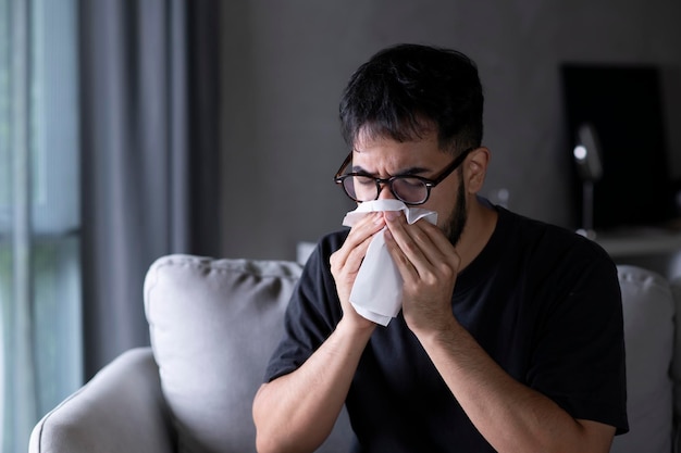 Asian man sick and sneeze with tissue paper on sofa at home Male blowing nose suffering from cold Cold and fever concept