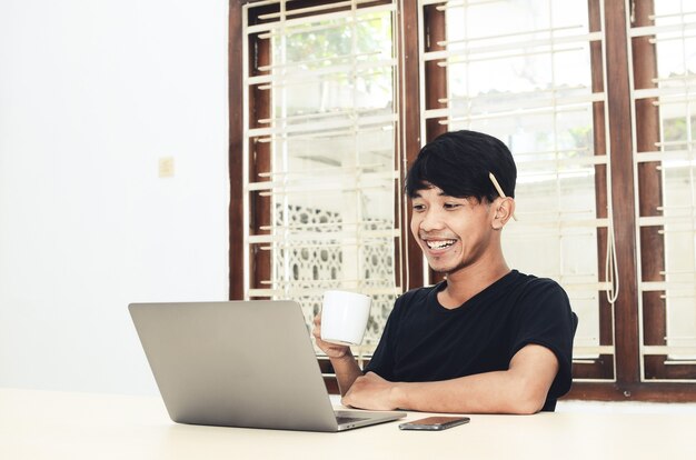 Asian man sits in front of a laptop drinking coffee