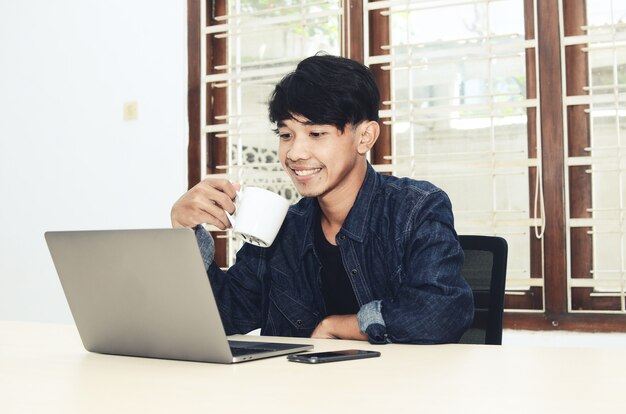 Asian man sits in front of a laptop drinking coffee