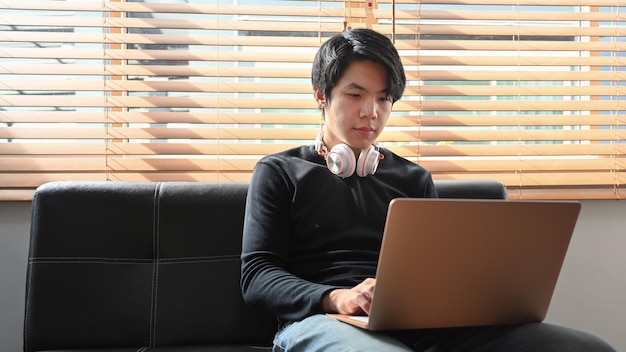ソファに座ってラップトップコンピューターでインターネットサーフィンをしているアジア人男性