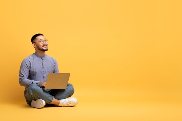 Asian Man Sitting On Floor With Laptop And Looking At Copy Space