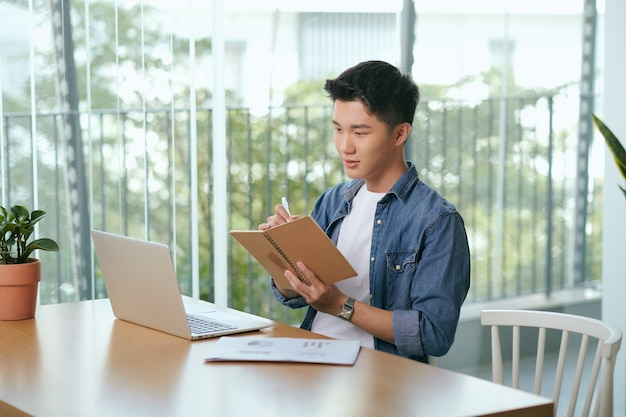 Asian man sitting in front of laptop and writing notes