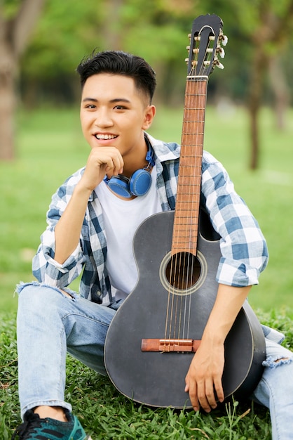 Asian man sitting on green grass with guitar and smiling at camera outdoors