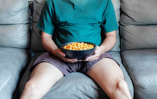 Foto uomo asiatico seduto sul divano con in mano una ciotola di popcorn