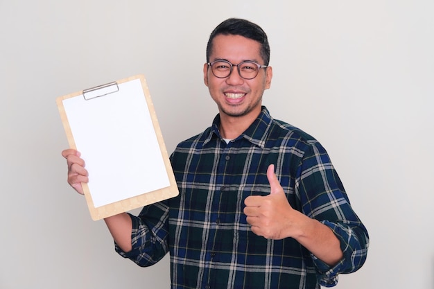 Photo asian man smiling and give thumb up while holding empty paper on clipboard