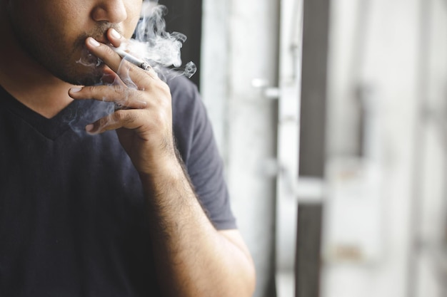 Foto uomo asiatico che fuma una sigaretta accanto alla finestra sfondo scuro concetto assistenza sanitaria