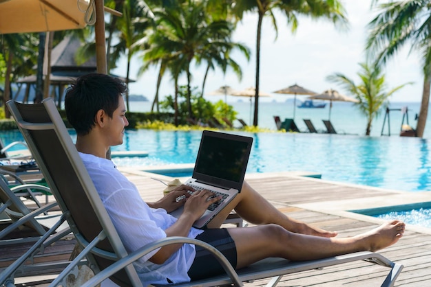 L'uomo asiatico ha trascorso le sue vacanze estive lavorando sul suo laptop su una sedia vicino alla piscina in un hotel resort