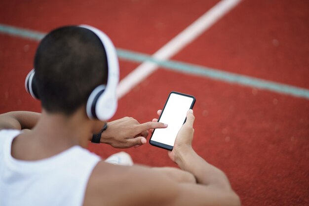 Photo asian man sportsman fit body wearing white headphone and using mobile phone on white screen to listen music while resting or before jogging exercising at lane stadium sport healthy running concept