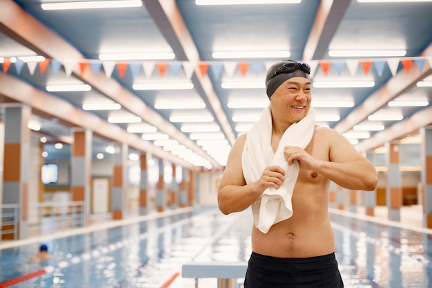 그의 어깨에 수건으로 실내 수영장에 서 있는 아시아 남자