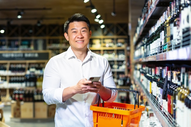 Asian man in supermarket uses phone and app to buy and choose wine in store looks at camera and smiles