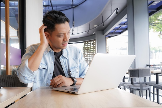 Asian man think and look for ideas with blue shirt using laptop and mobile phone in coffee shop