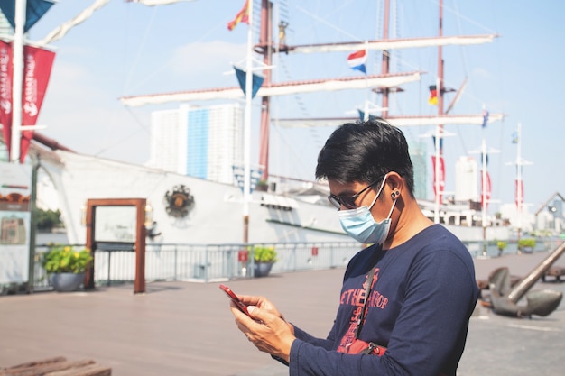 Turista asiatico dell'uomo che indossa la maschera per il viso utilizzando il telefono cellulare a bangkok posizione di viaggio