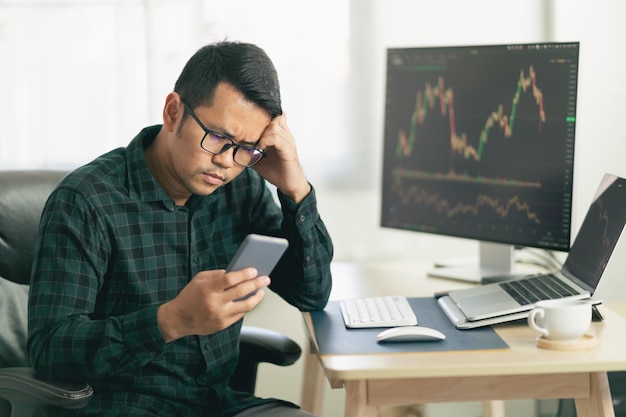 Asian man trader unhappy and serious sitting at home office in\
front of monitors with cryptocurrency graph holding smartphone\
monitoring cryptocurrency price