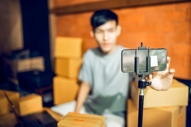 An asian man uses a mobile phone to take orders and display\
items in a box that records live streaming video online at the\
store small business owner asian online market delivery\
concept