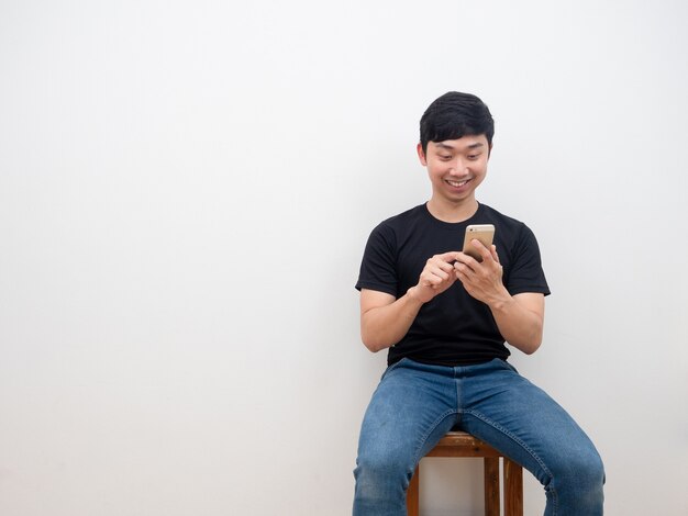 Uomo asiatico che utilizza lo smartphone in mano seduto su una sedia con un sorriso felice sullo sfondo del muro bianco