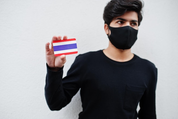 아시아 남성은 얼굴 마스크를 쓴 검은색 옷을 입고 흰색 배경 코로나바이러스 국가 개념에 격리된 태국 국기를 손에 들고 있습니다.