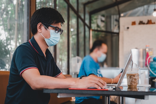 アジア人男性はコーヒーショップでラップトップコンピューターで作業しているフェイスマスクを着用し、他から社会的な距離を保ちます
