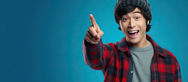 Asian man wearing beanie hat and red plaid flannel shirt energetically points upwards with finger on blue background