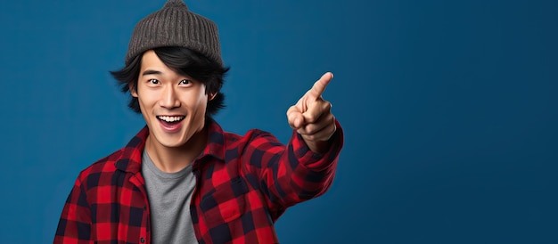 Asian man wearing beanie hat and red plaid flannel shirt energetically points upwards with finger on blue background