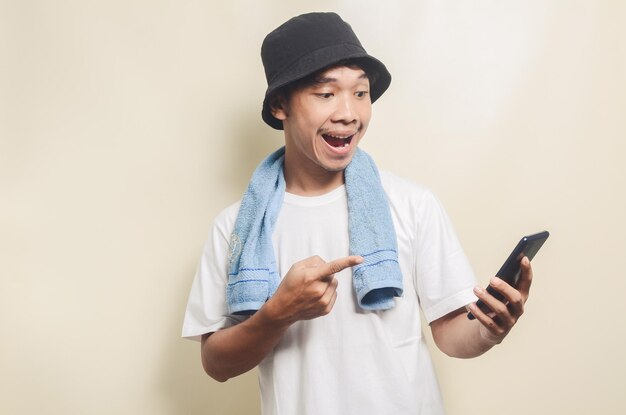Asian man wearing bright white tshirt in black hat with blue towel shocked to see his phone on isolated background