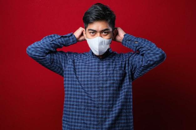 Asian man wearing casual shirt putting on a face mask with one hand putting the elastic band on an ear over red background