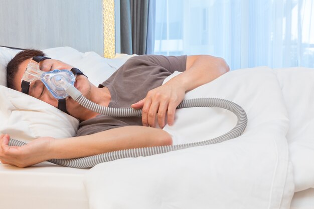 アジア人、CPAPマスクを着用し、エアホースに接続し、ベッドルームでベッドで寝る