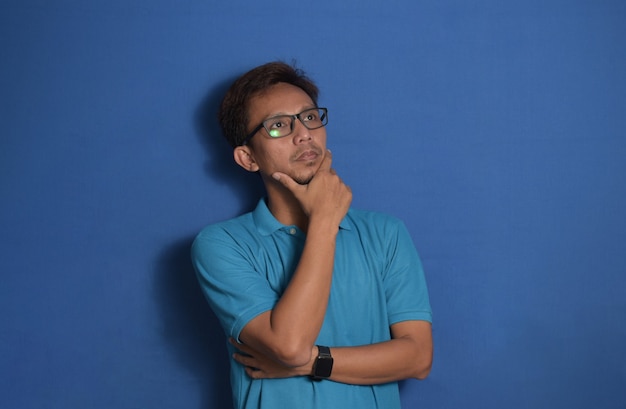 Asian man wearing glasses thinking and looking up over on blue background with hand on chin