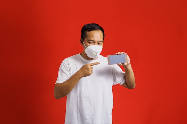 Asian man wearing medical mask pointing and holding smart phone with blank screen on red background