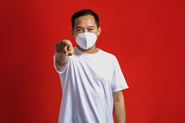 Азиатский мужчина в медицинской маске указывает и смотрит вперед на красном фоне