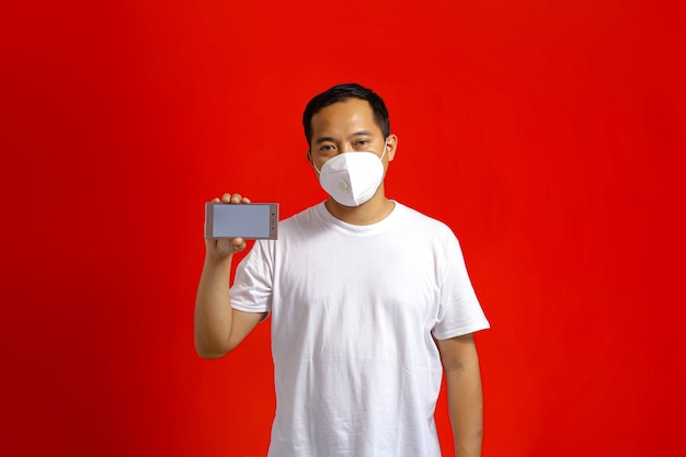 빨간색 배경에 빈 화면이 있는 스마트폰을 보여주는 의료용 마스크를 쓴 아시아 남자