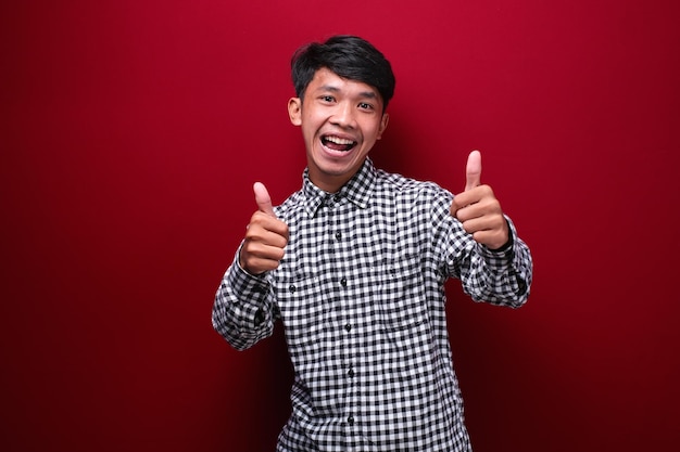 Asian man wearing plaid shirt with happy expression showing his two thumbs up on red background