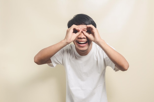 Asian man wearing white tshirt with gesture pretending to make binoculars with his hand on isolated background