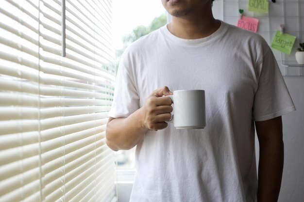 モックアップのための白いマグカップを保持している白いTシャツのアジア人男性