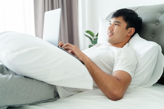 Foto uomo asiatico in maglietta bianca sdraiato sul letto utilizzando il computer portatile in camera da letto