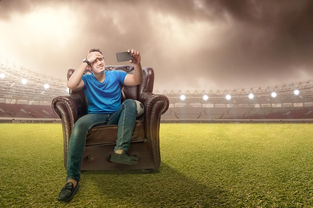 Asian man with the ball sitting on the couch with a disappointed expression watching a football match from a mobile phone