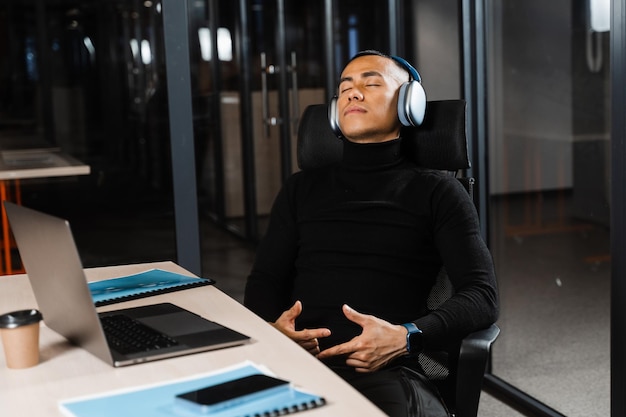 Asian man with closed eyes listening to relaxing music at work at laptop Break at workplace for rest from overtime work