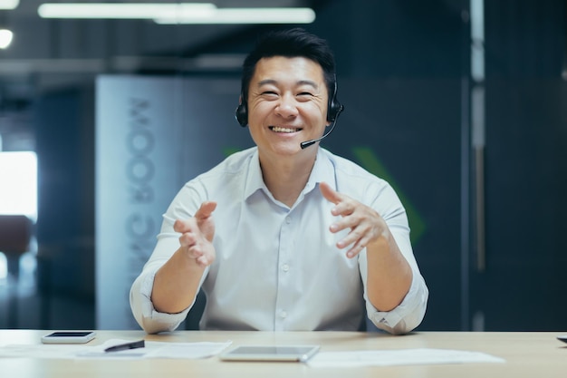 Uomo asiatico con l'auricolare che parla durante la videochiamata esaminando la webcam che sorride allegramente e