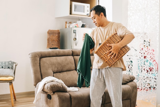 Asian man with laundry basket picking up dirty shirts from sofa at living room