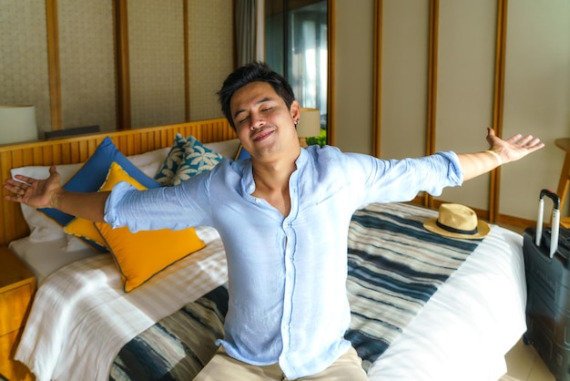 Asian man with a suitcase lay down on a bed in his room in resort after his arrival during his summer vacation holiday travel.