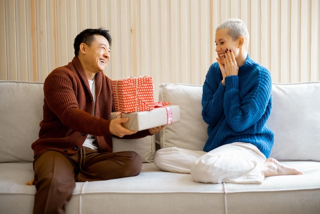 Asian man and young caucasian woman giving gifts each other. Concept of holiday, event and birthday. Idea of family relationship. Happy smiling couple sitting on sofa. Interior of modern apartment