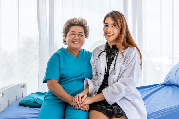 Asian medical doctor holding senior elderly woman female patient hand sitting on bed to comfort and motivate looking at camera