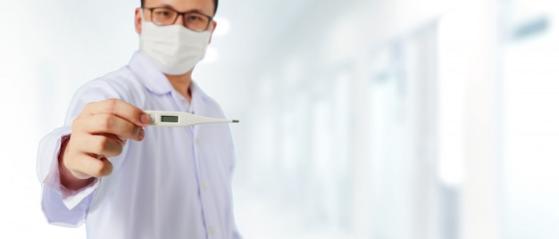 Asian medical male Doctor wear a mask to prevent Coronavirus 2019 and standing and raising hand to show thermometer in front of blurred hospital background.Concept of preventing infectious diseases.